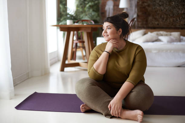 healthy active lifestyle, fitness, pilates, dieting and excess weight concept. charming barefooted plus size young woman in sportswear sitting cross legged on mat, going to practice yoga, smiling - healthy lifestyle dieting indoors lifestyles imagens e fotografias de stock