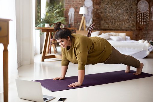 Attractive barefoot young overweight female doing plank on yoga mat while training indoors, watching online video via laptop. Sports, well being, technology and active healthy lifestyle concept