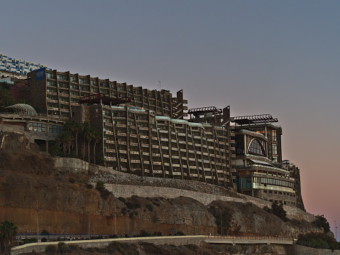 View of large ugly hotel building on the coast of holiday resort Puerto Rico de Gran Canaria, Canary Islands, Spain in the evening with road and rocks in winter season.
