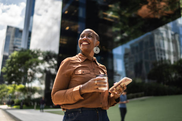 mujer de negocios sosteniendo un teléfono inteligente y mirando hacia otro lado al aire libre - desafío fotografías e imágenes de stock