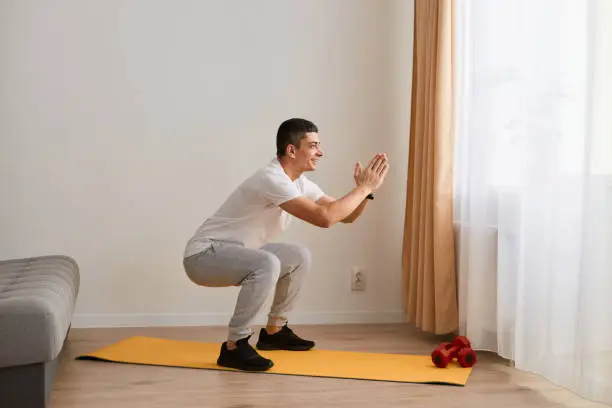 Side view portrait of man wearing sportswear doing sport exercises at home, doing split or squat exercise with stretched out hands, lower-body exercise.