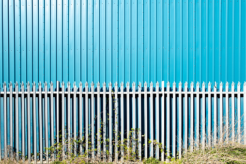 Abstract image of an newly built metal warehouse building seen behind a metallic fence. Nettles are seen in front of the fence.