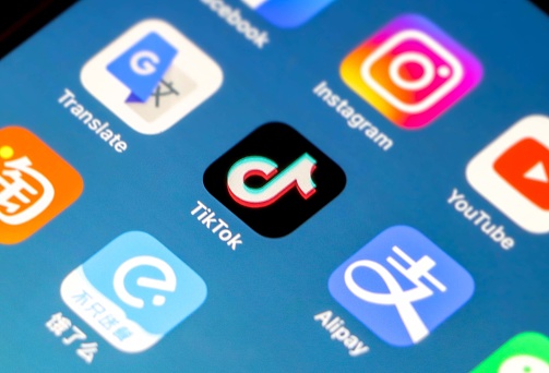 Chinese company Bytedance’s hugely popular short-form video social networking platform - Tik Tok's Application Icon in focus on iPhone smartphone screen, other apps (Alipay, Instagram etc) out of focus on blue gradient background wallpaper