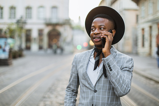 Stylish african american man in black hat having mobile conversation while standing on city street. Young male hipster walking and taking outdoors.