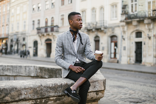 African american man in stylish suit sitting on old fountain with cup of coffee. Young male hipster enjoying hot beverage while resting on city street.