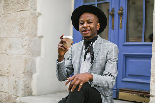 Positive african american guy in black hat and stylish suit smiling and looking at camera while sitting with cup of coffee outdoors. Concept of youth, lifestyles and leisure activity.