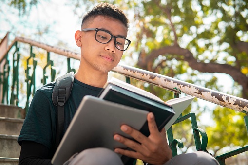 Outdoor low angle image of confident Asian teenager, student sitting on stairs and doing study in the college campus day time.