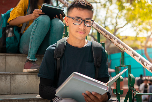 Imagen al aire libre de un adolescente asiático, un estudiante sentado en las escaleras y estudiando en el campus universitario durante el día. Está mirando a la cámara con una sonrisa confiada. photo