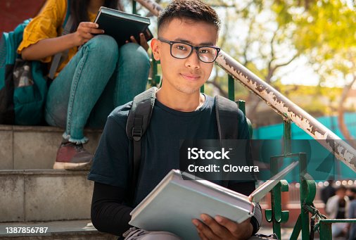 istock Outdoor image of Asian teenager, student sitting on stairs and doing study in the college campus day time. He is looking at the camera with a confident smile. 1387856787