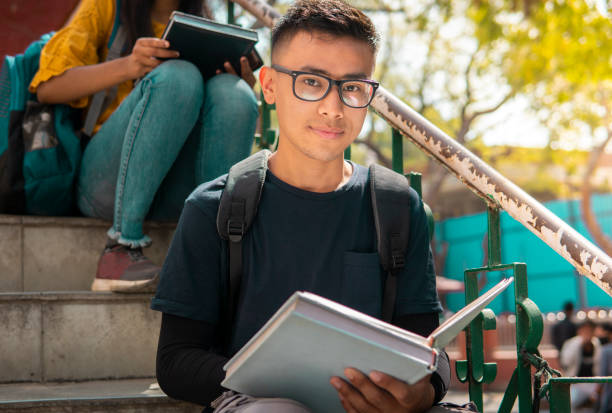 imagem ao ar livre de adolescentes asiáticos, estudante sentado em escadas e estudando no dia do campus da faculdade. ele está olhando para a câmera com um sorriso confiante. - aluno do ensino médio - fotografias e filmes do acervo