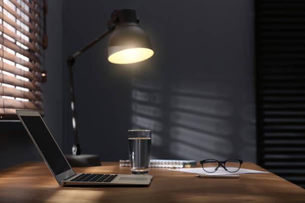 istock Modern laptop and lamp on table near window in office. Space for text 1387857923