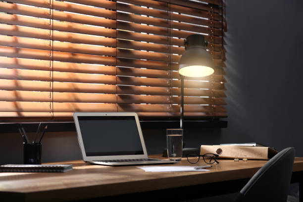 istock Modern laptop and lamp on table near window in office 1387858910