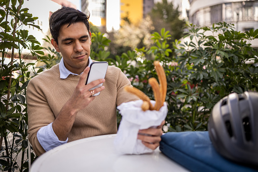 Young businessman using mobile phone. He is sitting at cafe and eating churros