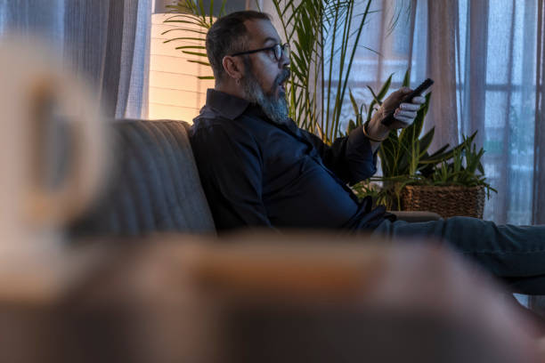 istock Adult man sitting on sofa changing tv channels with remote. 1387872628