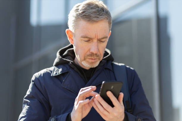Outdoor portrait of a 50 year old man with a beard in a blue jacket with a mobile phone stock photo