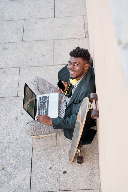 istock Young businessman sitting outdoors at a wall working on laptop looking at camera with smartphone 1387877730