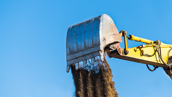 Close-up of yellow digger arm metal bucket when dumping brown dirt. Working hydraulic earth mover detail