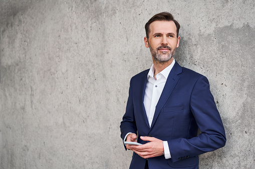 Portrait of confident businessman standing against concrete wall holding smartphone looking away