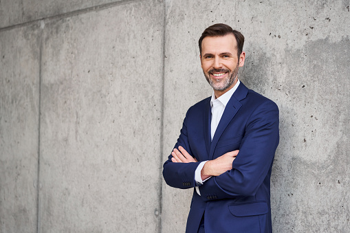 Portrait of smiling successful businessman in suit standing against concrete wall