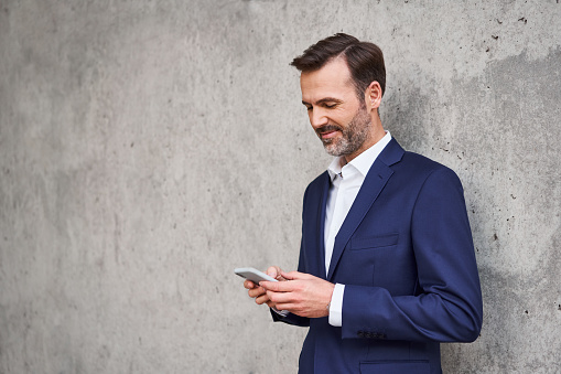 Portrait of smiling businessman in suit using mobile phone standing against concrete wall