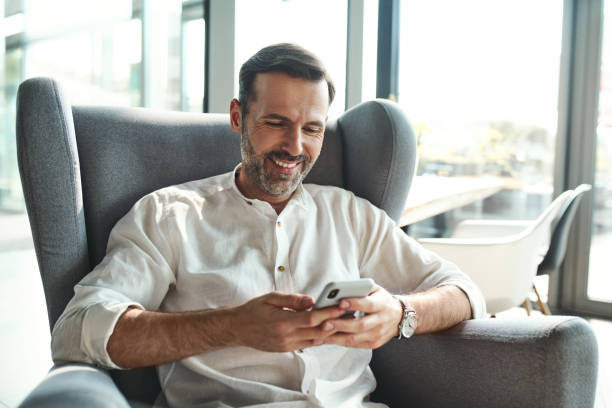 Smiling mid adult man sitting on armchair in modern house using smartphone stock photo