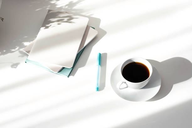 istock Black coffee cup, stack of notebooks or planners on white table with interesting shadows. Scandinavian style, stationary still life in the morning rays of sun. 1387933154