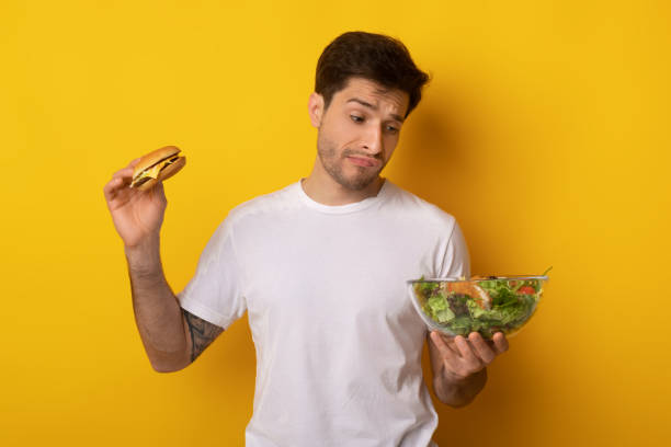 Portrait of Funny Guy Holding Burger And Vegetable Salad Healthy Or Unhealthy Food Concept. Puzzled Confused Man Holding Burger And Looking At Bowl With Vegetable Salad. Hungry Guy Choosing What To Eat, Thinking Isolated On Yellow Orange Studio Background EATING HEALTHY stock pictures, royalty-free photos & images