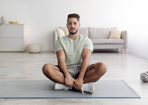 jeune arabe équilibré assis les jambes croisées sur un tapis de sport, méditant ou pratiquant le yoga à la maison, pleine longueur - men yoga meditating cross legged photos et images de collection