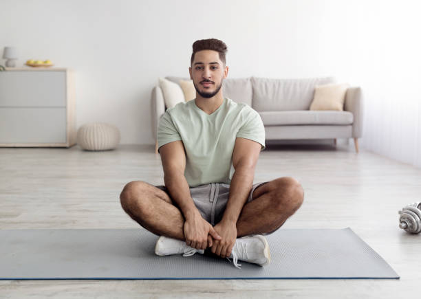 jovem árabe equilibrado sentado de pernas cruzadas em tapete esportivo, meditando ou praticando yoga em casa, comprimento completo - men yoga meditating cross legged - fotografias e filmes do acervo