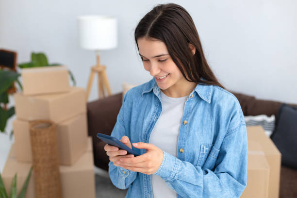 Moving to a new house, rental housing. Happy young caucasian woman using a mobile phone to search and order a transportation service and movers to move to a new home stock photo