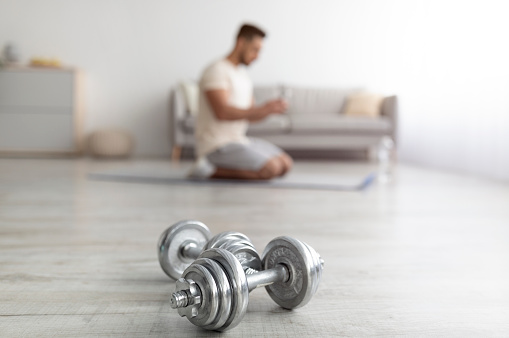 Millennial Arab guy exercising at home during covid lockdown, selective focus on dumbbells. Young middle Eastern man having domestic training, doing bodybuilding workout indoors, copy space