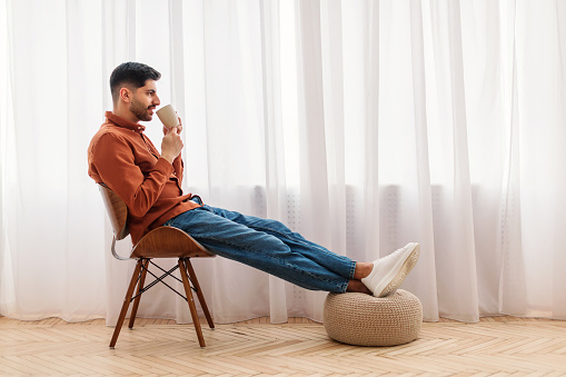 Rest Concept. Happy Arab guy drinking coffee sitting on chair in living room. Casual man relaxing, enjoying weekend free time or break from work, free copy space, profile side view, full body length