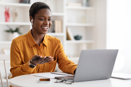 Attractive young african american woman having video conference with clients or business partners, sitting at workdesk at office, using laptop and earbuds, gesturing and smiling, copy space