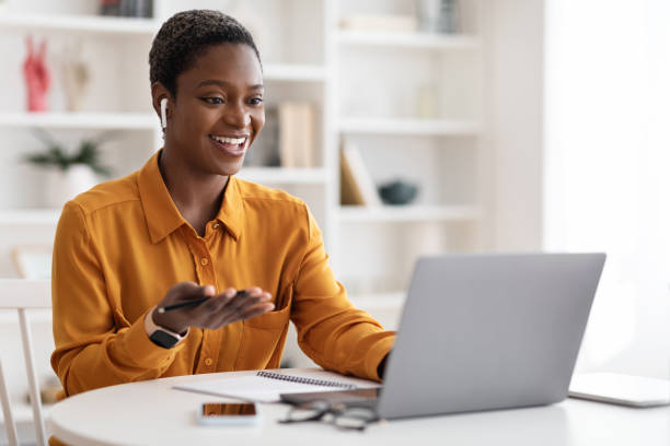 Young black woman having video conference with clients, copy space Attractive young african american woman having video conference with clients or business partners, sitting at workdesk at office, using laptop and earbuds, gesturing and smiling, copy space video conference stock pictures, royalty-free photos & images
