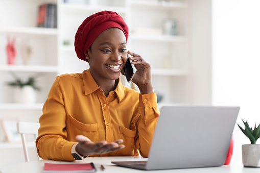 Positive young muslim black lady with red turban on her head and casual outfit working on laptop at office, looking at computer screen and talking on cell phone with business partner, copy space