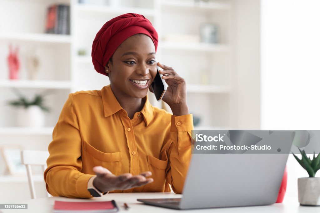 Positive muslim black lady working on laptop at office Positive young muslim black lady with red turban on her head and casual outfit working on laptop at office, looking at computer screen and talking on cell phone with business partner, copy space Using Phone Stock Photo