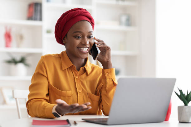 Positive muslim black lady working on laptop at office Positive young muslim black lady with red turban on her head and casual outfit working on laptop at office, looking at computer screen and talking on cell phone with business partner, copy space using phone stock pictures, royalty-free photos & images