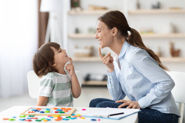 Speech training for kids. Professional woman training with little boy at cabinet, teaching right articulation exercises stock photo