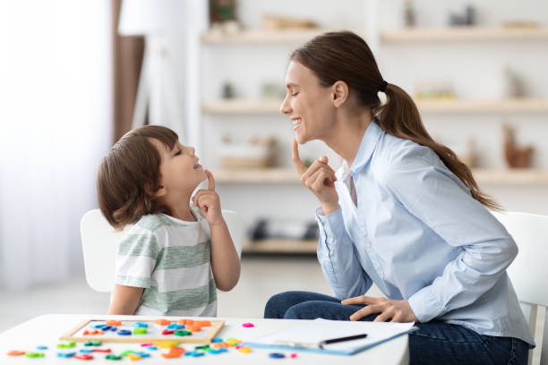 Speech training for kids. Professional woman training with little boy at cabinet, teaching right articulation exercises Speech training for kids. Professional woman specialist training with little boy at cabinet, teaching him right articulation exercises, side view autism stock pictures, royalty-free photos & images