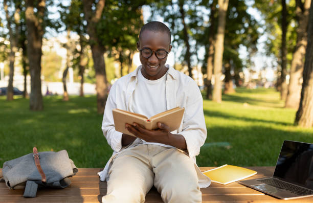 istock Male african american student reading book outdoors, preparing for classes, sitting in park or university campus 1387961349