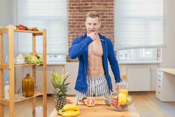 молодой красивый атлетичный мужчина ест яблоко на кухне дома, готовит свежевыжатый сок и фруктовый салат, смотрит в камеру - fruit salad fruit healthy eating making стоковые фото и изображения