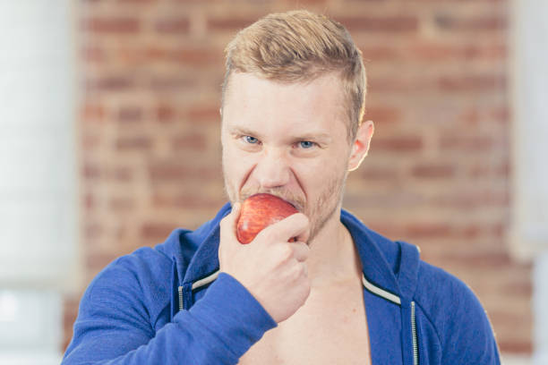 фото крупным планом. портрет молодого красивого атлетического атлетического мужчины, поедающего яблоко на кухне дома, д�елающего свежевыжа - fruit salad fruit healthy eating making стоковые фото и изображения