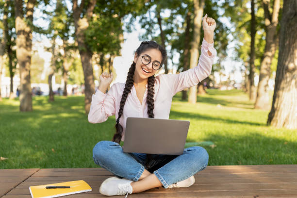 istock Overjoyed indian female student with laptop computer gesturing YES outdoors, sitting in university campus 1387963670