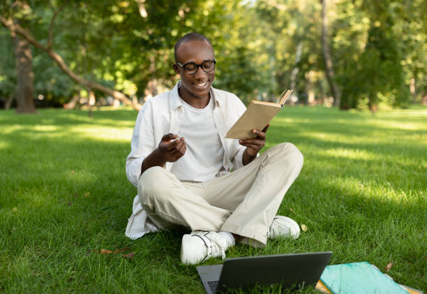 istock Exam study concept. Happy african american student guy reading book and using laptop computer outdoors 1387963718