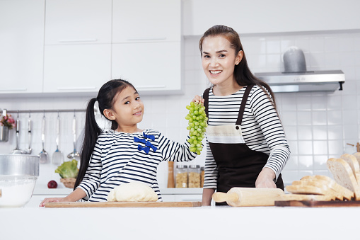 Smiling Asian mother and little asian girl child funny with grape fruit and baking bakery together in kitchen. Homemade pastry for bread. Family love and education for Homeschool Concept.