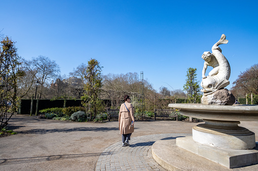 A woman walks past Boy and Dolphin Fountain at the Rose Garden of Hyde Park in City of Westminster, London. The marble monument was designed in 1862 by Alexander Munro