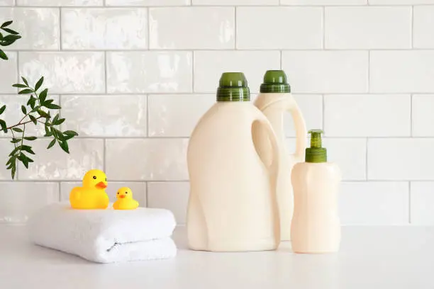 Photo of Eco friendly organic natural baby laundry detergent and soap gel bottle with branch of green leaves, towel and yellow duck on table in bathroom. Baby hygiene products packaging design, branding.