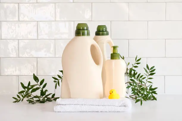 Photo of Eco friendly organic natural baby laundry detergent container and soap gel bottle with branch of green leaves, towel and yellow duck on table in bathroom.