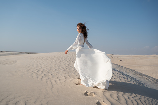 Young bride in white waving gown walking on a sand dune in desert