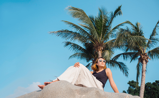 Asian woman look cool with short hair wearing sunglasses, black tank top and white trousers sitting on the rock at the beach under the coconut palm trees and blue sky background, summer style fashion.